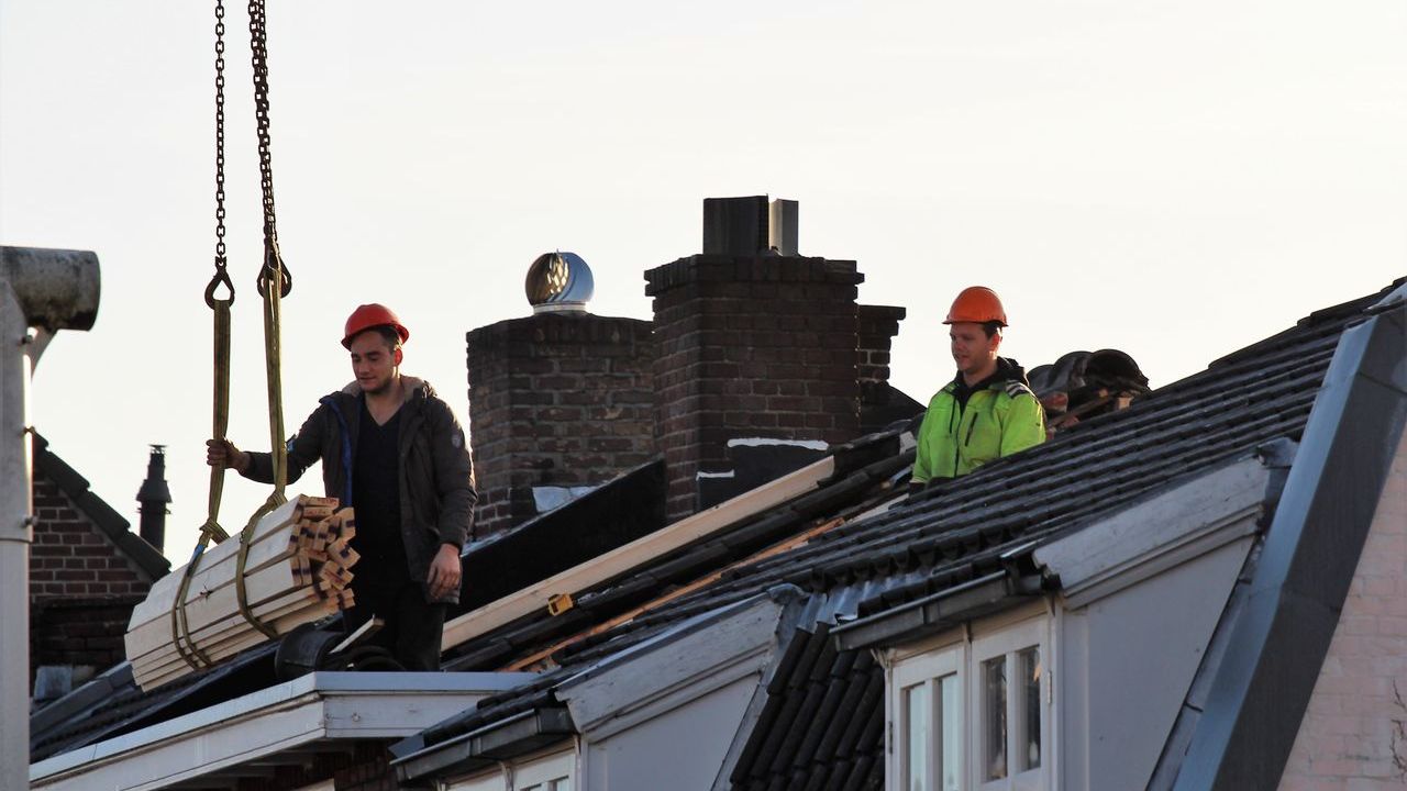 To håndværkere i orange sikkerhedshjelme løfter en tagbjælke på plads på en byggeplads.
